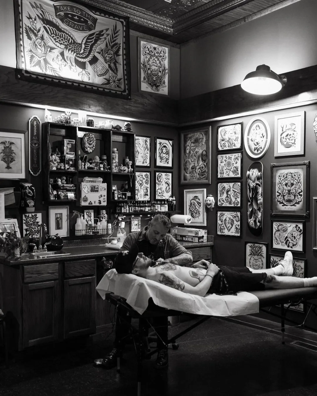 An artist tattooing a client's chest at a station walled with framed flash, under a hanging lamp
